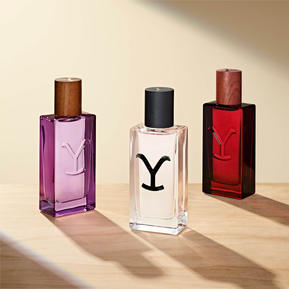 Three Yellowstone perfume bottles in purple, black, and red on a wooden surface with a beige background.