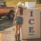 Woman in a cowboy hat leaning against an ice box next to a vintage truck on a rural road.