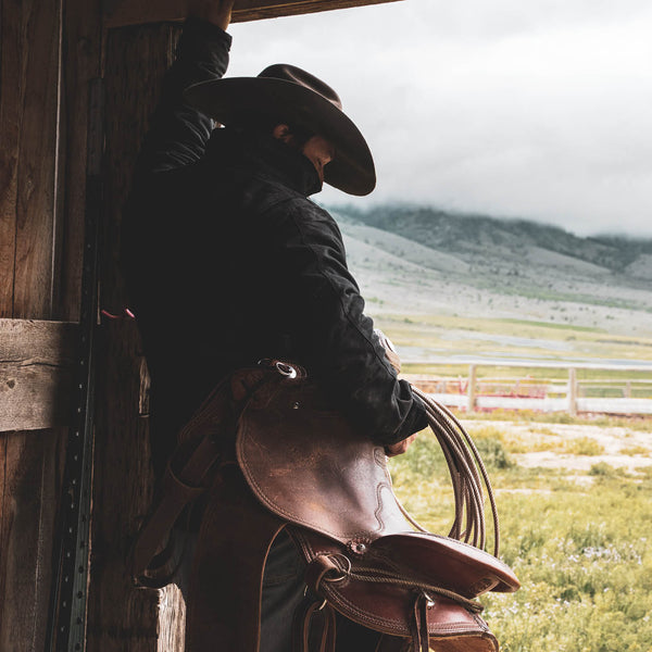 Person holding a leather saddle in a rustic setting with mountains in the background