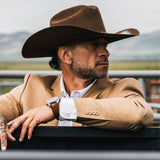Man wearing a cowboy hat and beige jacket sitting in a vehicle with a scenic background