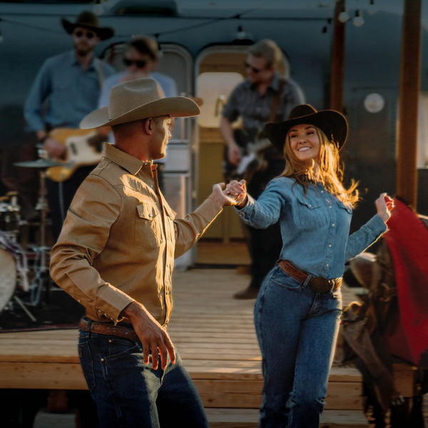 Man and woman in cowboy hats dancing on a wooden platform with a band in the background.