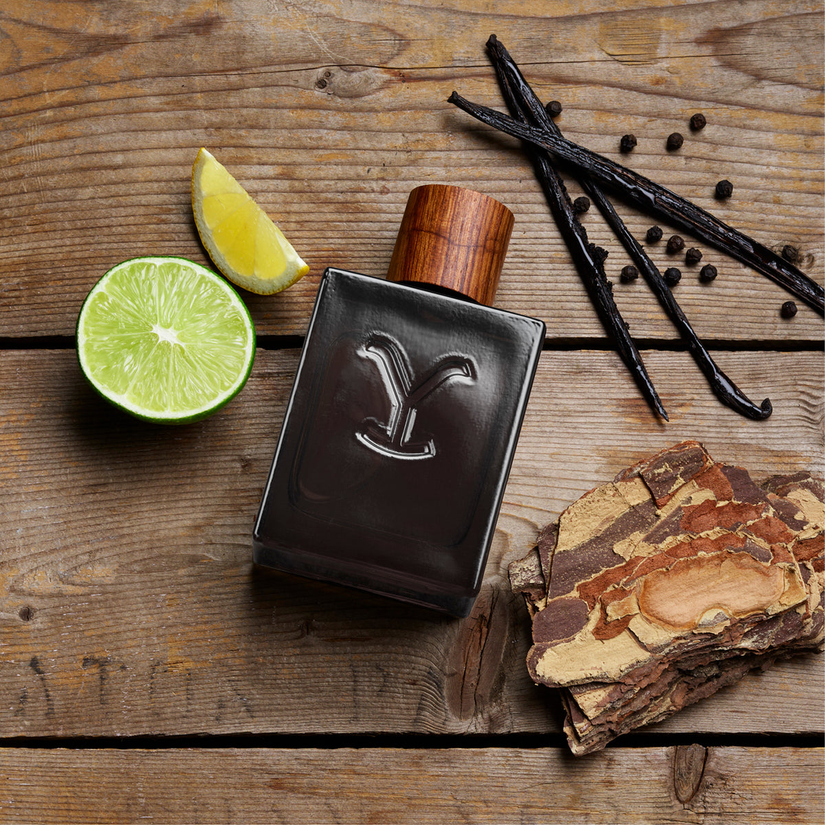 Perfume bottle with wooden cap on a wooden surface with lime, vanilla beans, and bark.