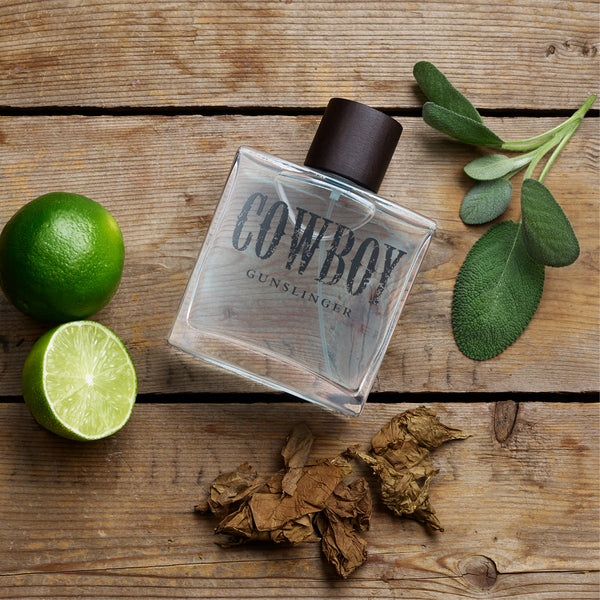 Perfume bottle labeled 'Cowboy Gunslinger' on a wooden surface with limes and tobacco leaves.