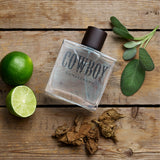 Perfume bottle labeled 'Cowboy Gunslinger' on a wooden surface with limes and tobacco leaves.