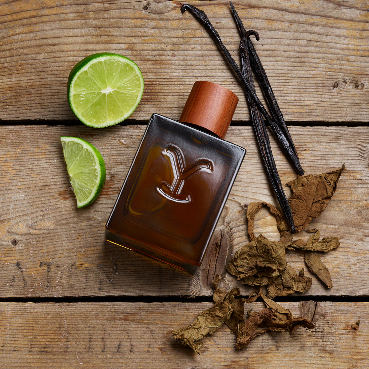 Perfume bottle with vanilla beans, lime slices, and dried leaves on a wooden surface